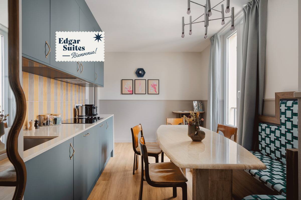 Een moderne keuken met een blauw eiland, houten stoelen en een eettafel. Er hangen foto's in lijsten aan de muur en er is een raam met gordijnen.