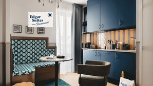 Een compacte kamer met een geruit bankje, een ronde tafel, een stoel en een kitchenette met kastjes en een aanrecht.