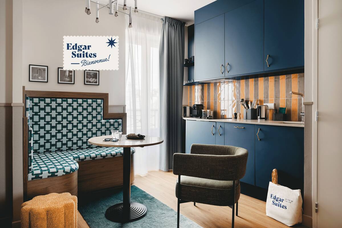 Een compacte kamer met een geruit bankje, een ronde tafel, een stoel en een kitchenette met kastjes en een aanrecht.