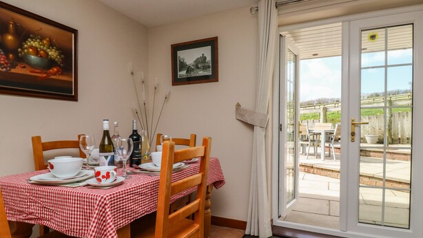A dining area with a checkered tablecloth, wooden chairs, and a table set for two. There are wine bottles and glasses on the table. - Otters Holt (South Molton)