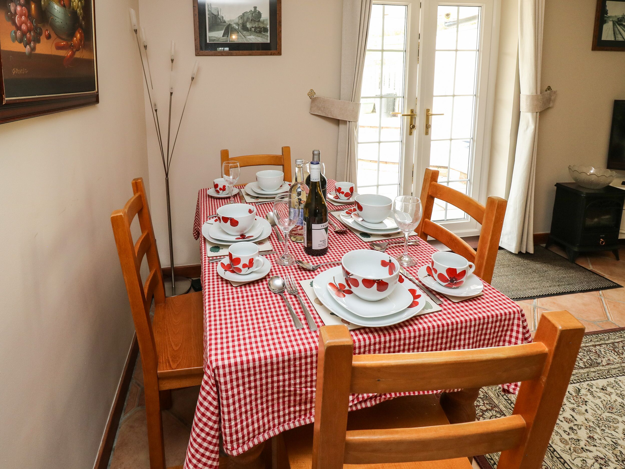 Une table à manger dressée avec de l’argenterie blanche, des assiettes et des bols, une nappe à motifs floraux rouges et des chaises en bois.