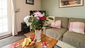 A living room with a sofa, a coffee table with fruit, a vase with flowers, a framed picture on the wall, and a radiator. - Otters Holt (South Molton)
