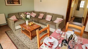A cozy living room with a sofa set, a coffee table with a vase of flowers, and a dining area with a checkered tablecloth, plates, and wine bottles. - Otters Holt (South Molton)