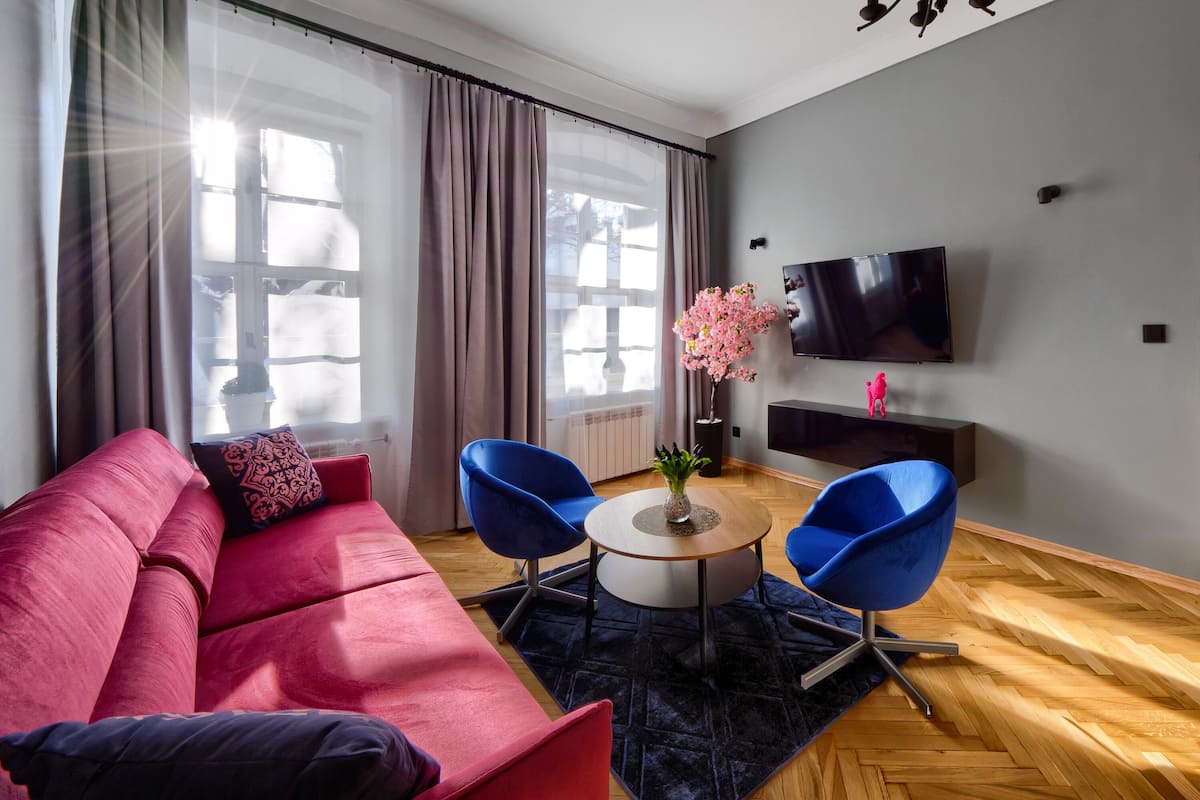 Ein modernes Wohnzimmer mit einem roten Sofa, zwei blauen Sesseln, einem kleinen runden Tisch und einem Fernseher an der Wand.