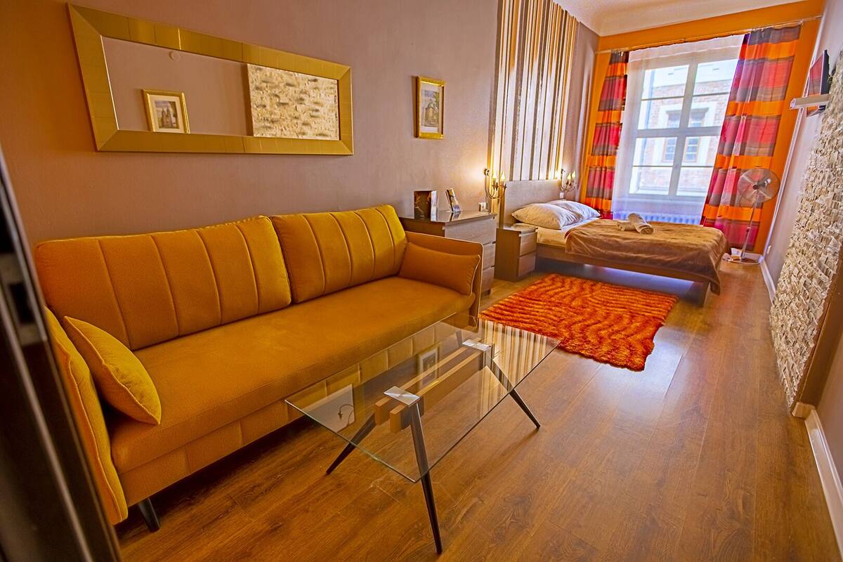 Ein modernes Wohnzimmer mit einer Couch, einem Glastisch, einem Bett mit gestreifter Decke und einem Fenster mit Vorhängen.