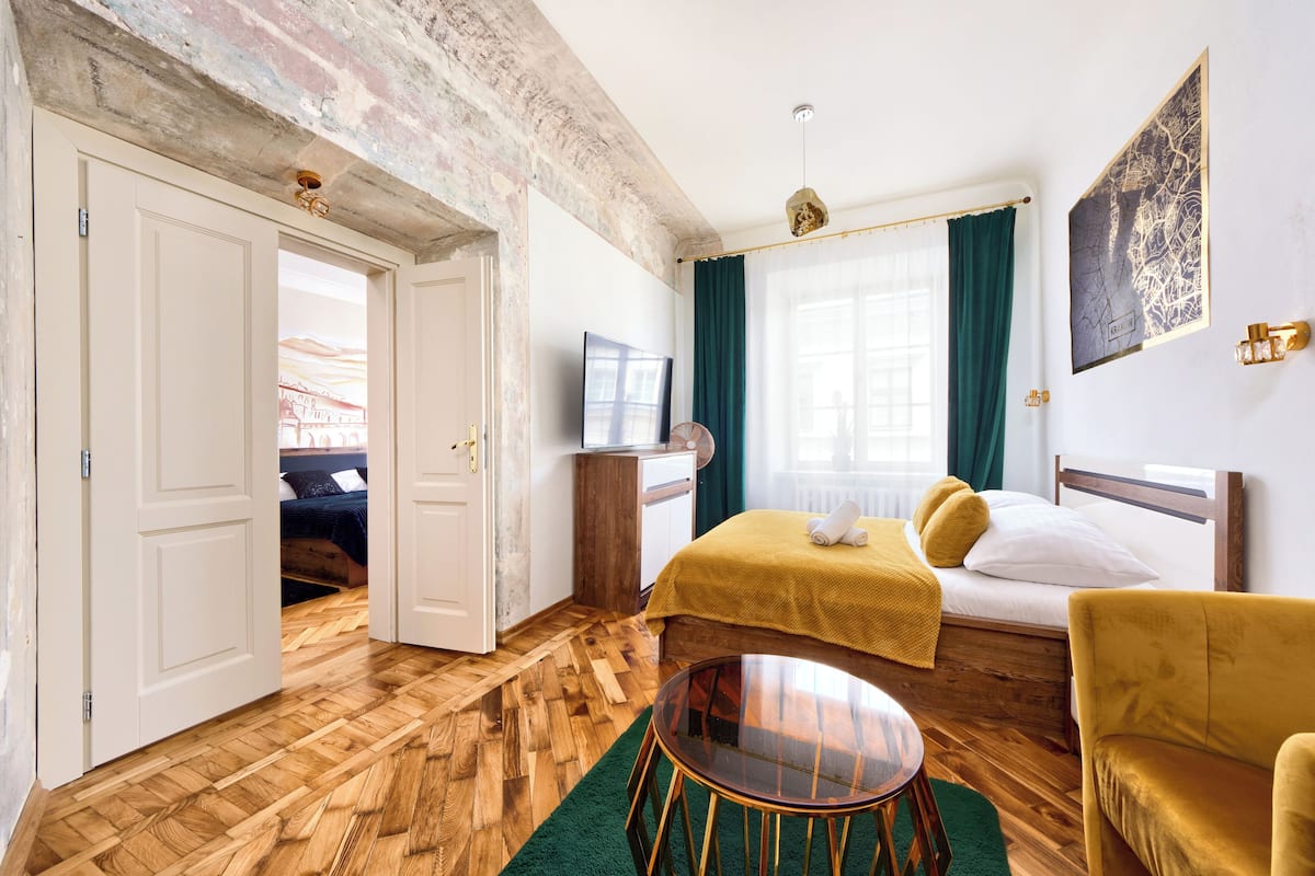Ein Hotelzimmer mit Holzboden, einem Bett mit gelber Decke, einem grünen Teppich, einem runden Tisch und einem Bild an der Wand.