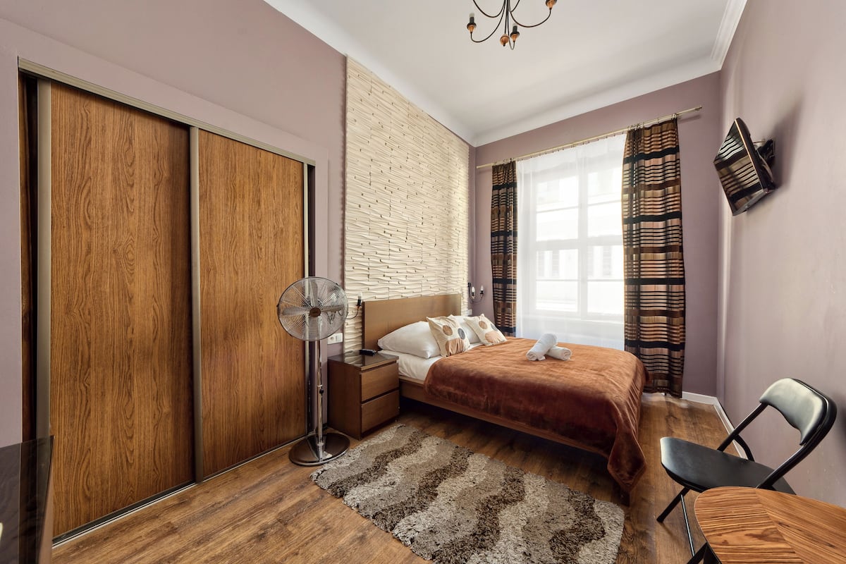 Ein Schlafzimmer mit Bett, Nachttisch, Ventilator und einem Holzschrank.