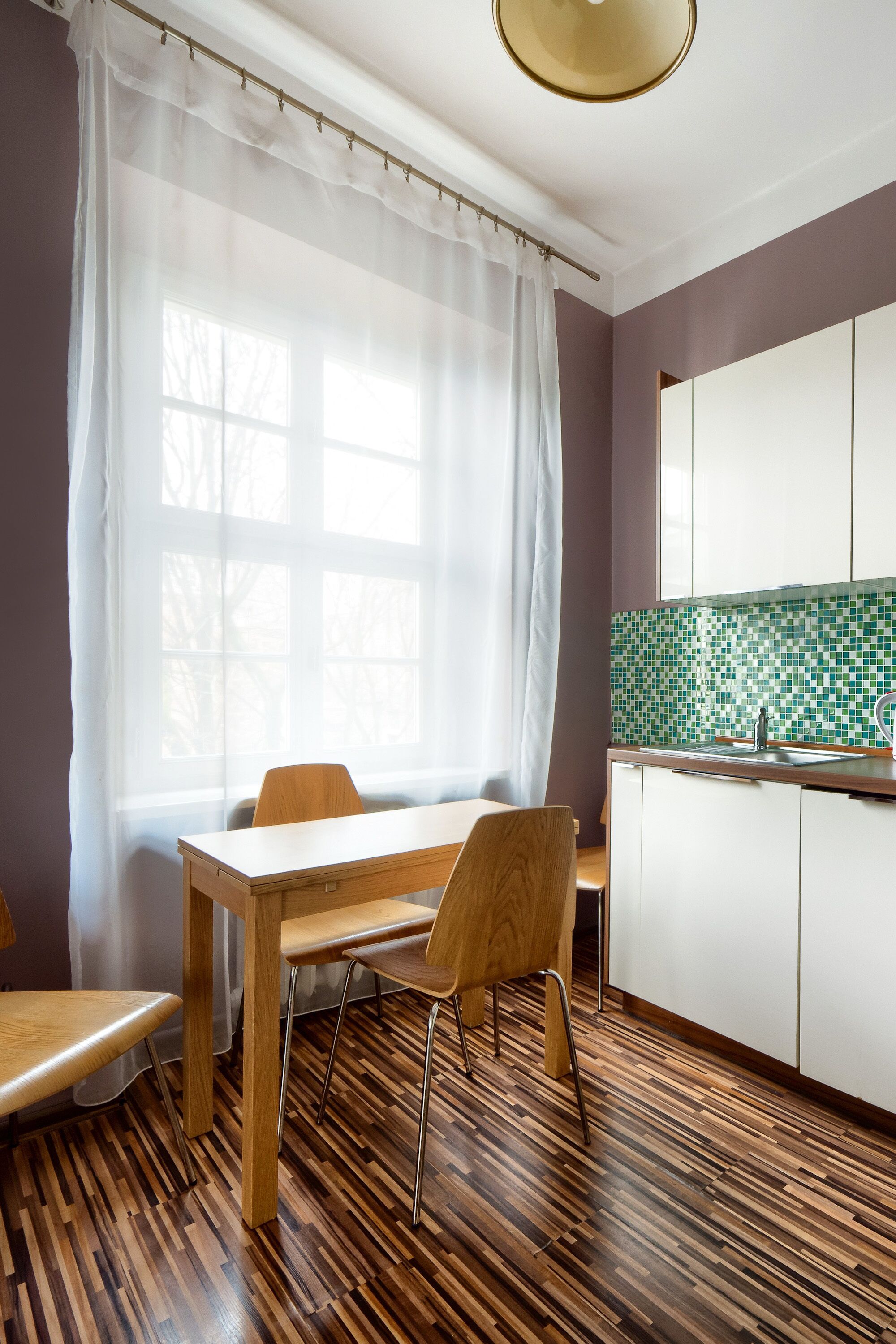 A kitchen with a wooden dining table, chairs, a window with sheer curtains, and a tiled backsplash.
