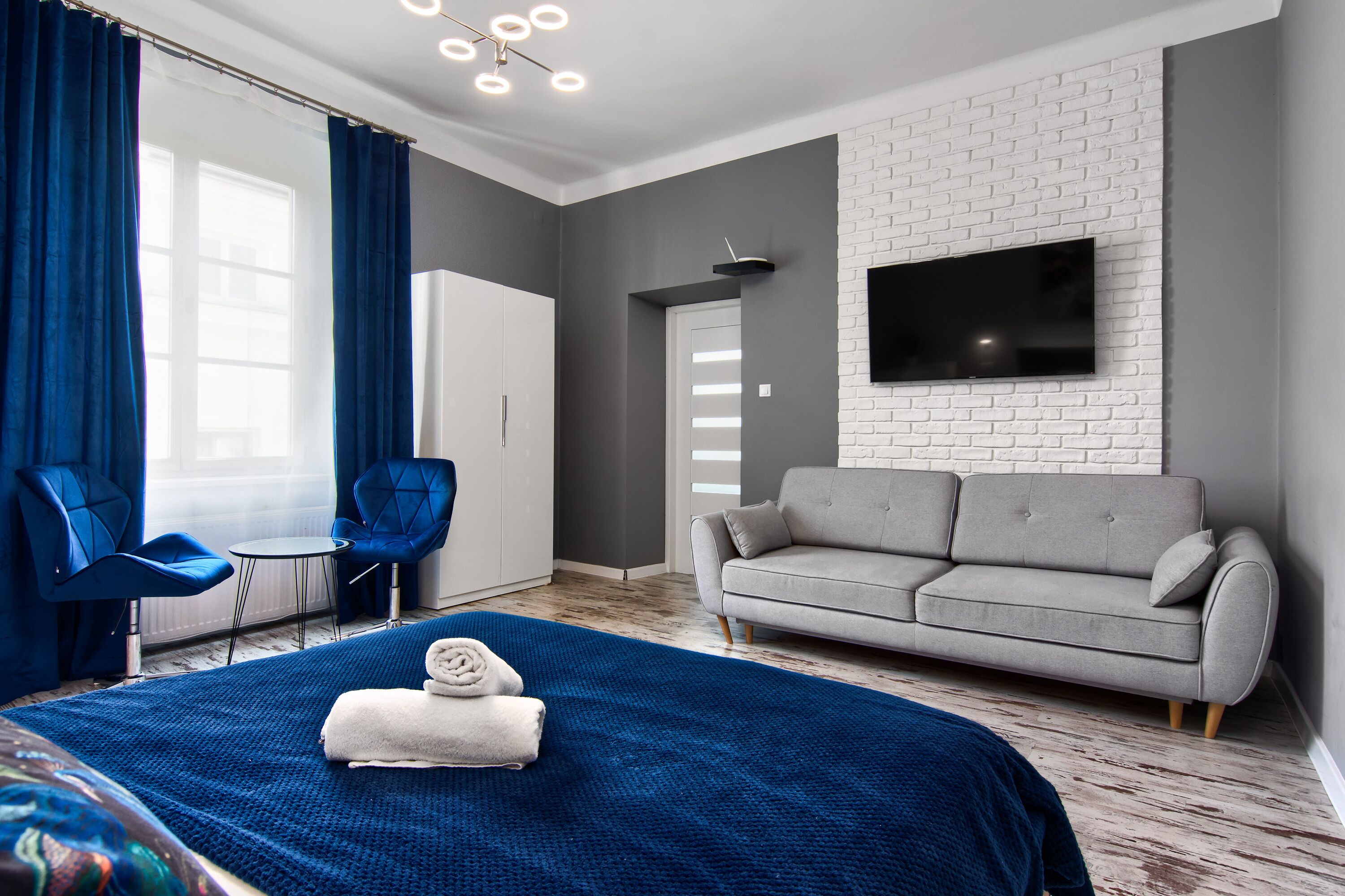 Una habitación de hotel moderna con una cama grande, un sofá gris, dos sillones azules, un televisor de pantalla plana montado en una pared de ladrillo blanco y una mesita pequeña.