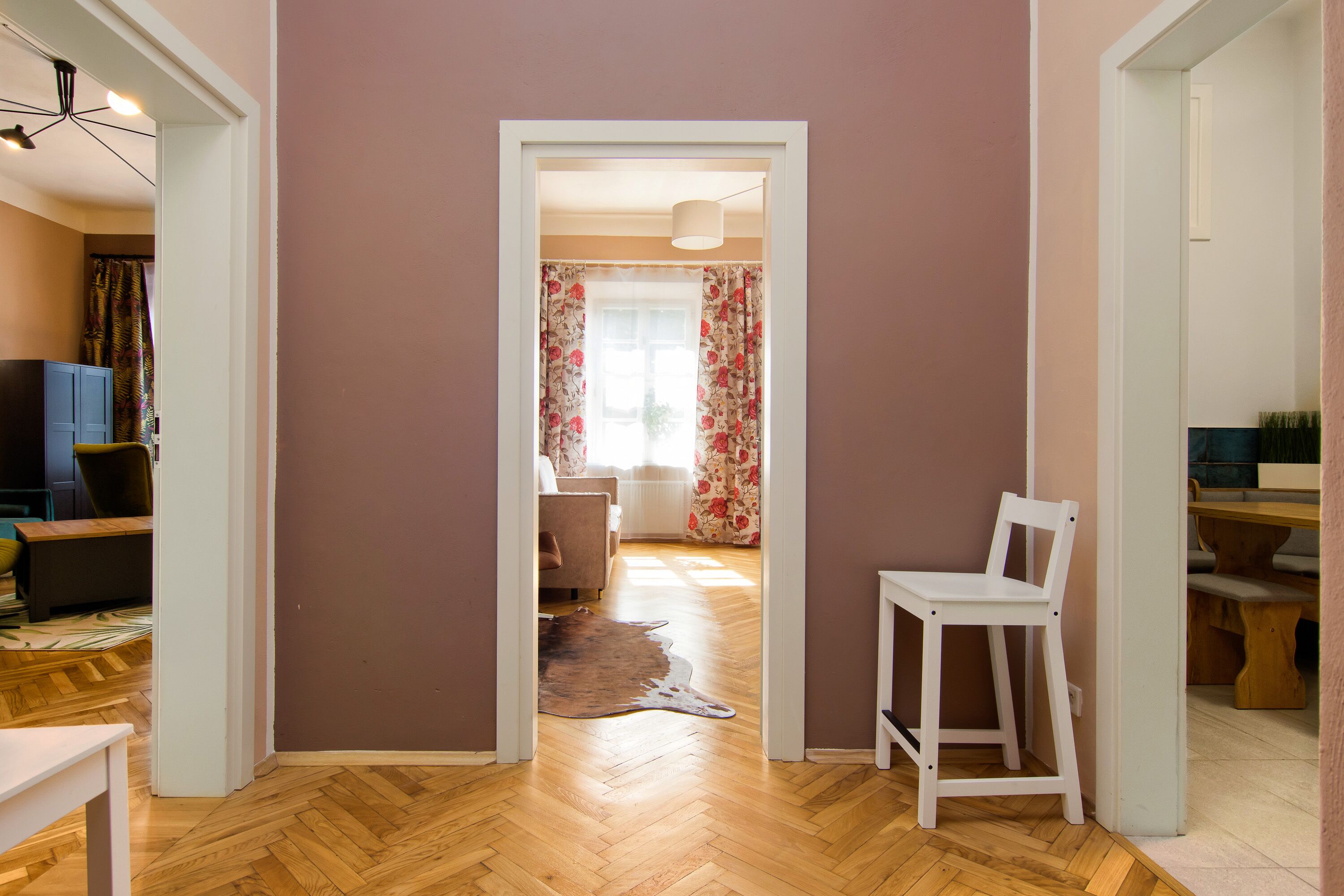 A hallway with a wooden floor, a white chair, and a door leading to a room with a bed and curtains.