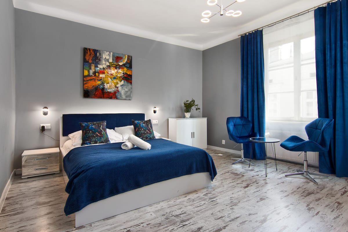 Ein modernes Schlafzimmer mit einem großen Bett, einem Nachttisch, einer kleinen Kommode und einem Stuhl an einem Fenster mit blauen Vorhängen.