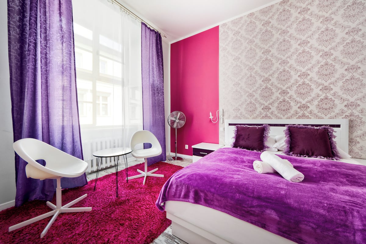 Ein modernes Schlafzimmer mit einem großen Bett, violetter Bettwäsche, einem weißen Stuhl und einem kleinen Tisch.