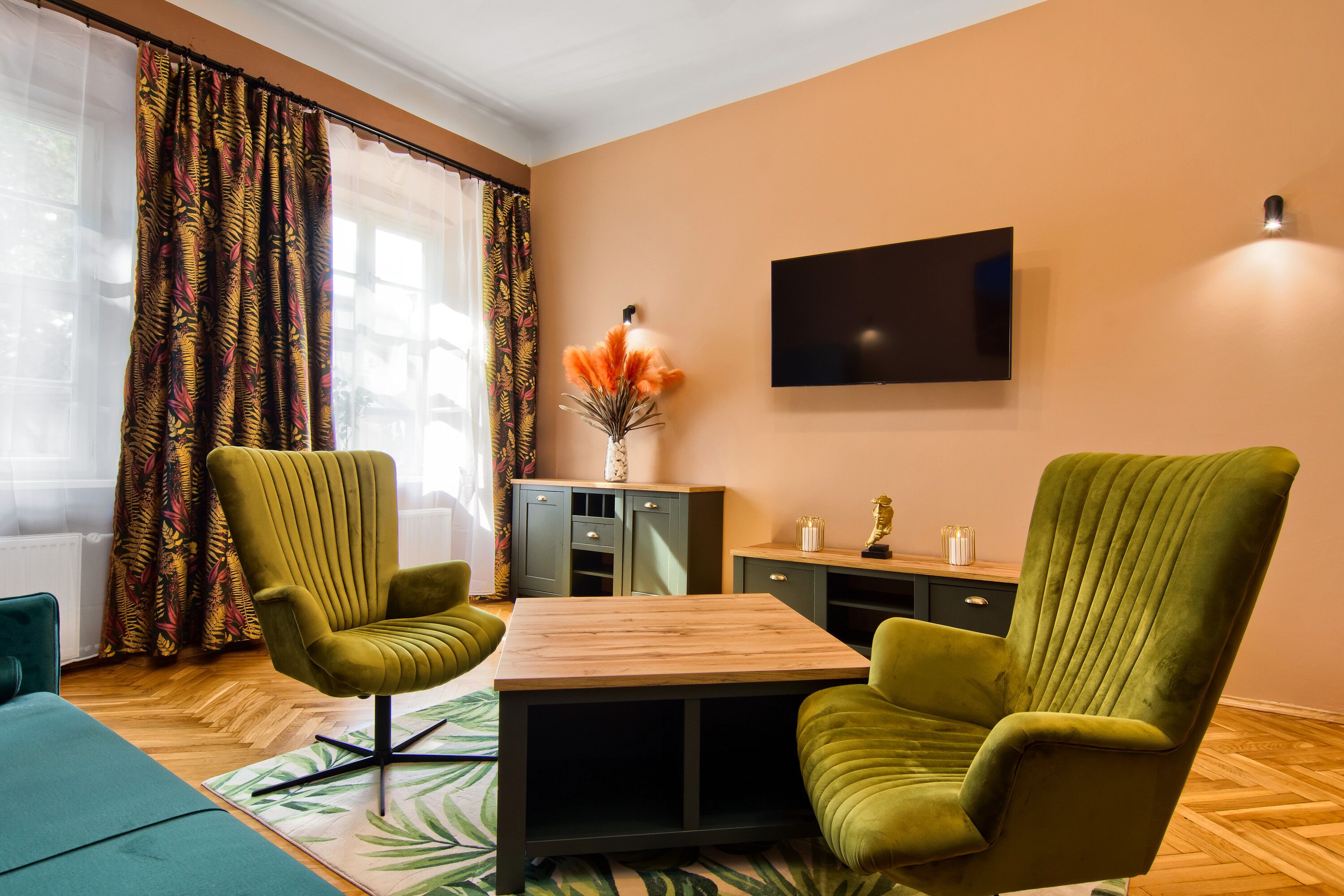 Une pièce avec deux fauteuils verts, une table en bois, un téléviseur fixé au mur et un meuble contenant des objets décoratifs.