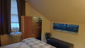 Un dormitorio con cama, mesita de noche, ventana con cortinas, armario de madera, banco y un cuadro en la pared.