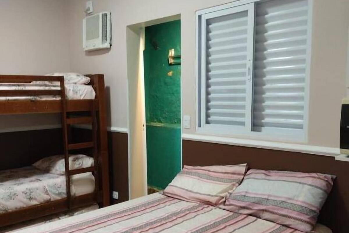 Une chambre avec un lit superposé, un lit simple, une fenêtre avec des stores et une porte verte.