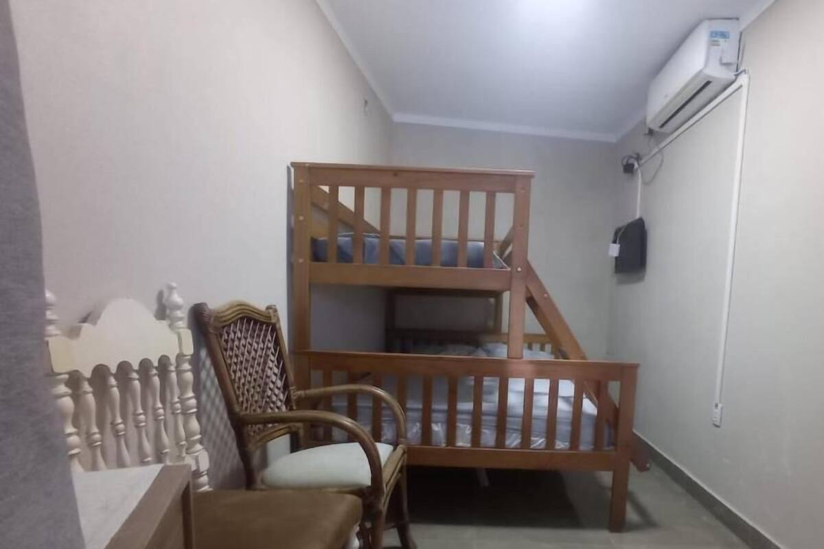 Un lit superposé avec une structure en bois et un lit simple en dessous. On y trouve une chaise en bois avec un coussin, une petite table et une clôture décorative. Un appareil de climatisation est fixé au mur.