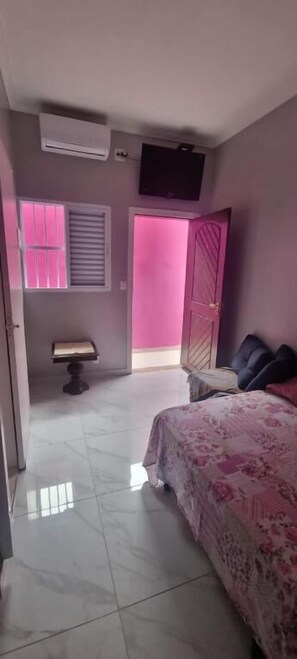 Een slaapkamer met een bed, nachtkastje, raam met zonwering, deur en een airconditioner aan het plafond.