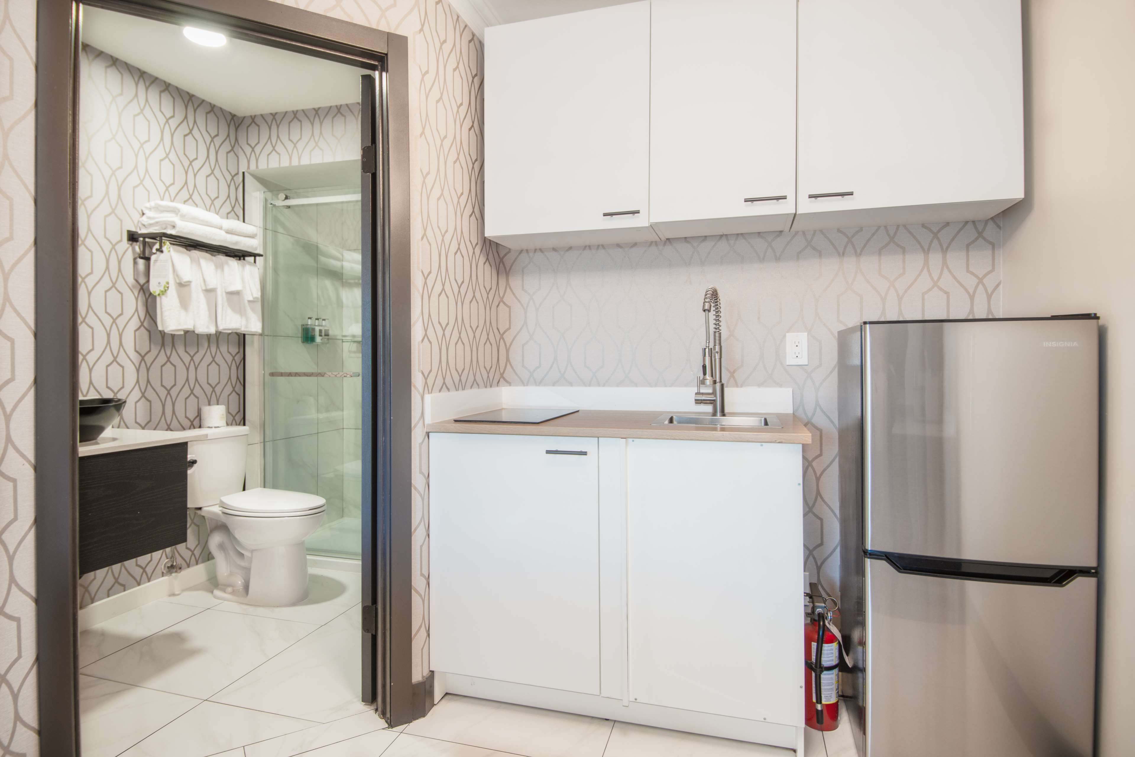 Un baño moderno con inodoro, lavamanos y ducha. Hay un armario blanco sobre el lavamanos y un refrigerador de acero inoxidable a la derecha.