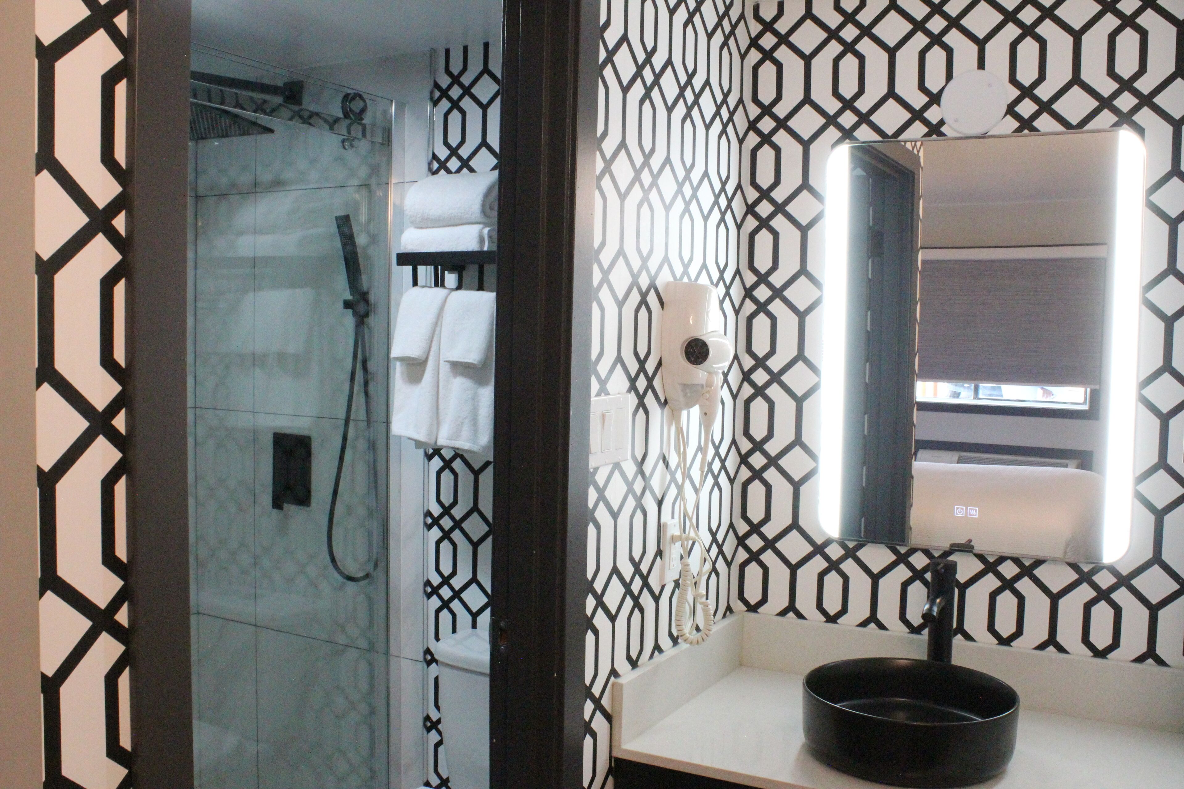 A bathroom with a glass shower enclosure, a white sink with a black basin, and a mirror reflecting a towel rack and a window.