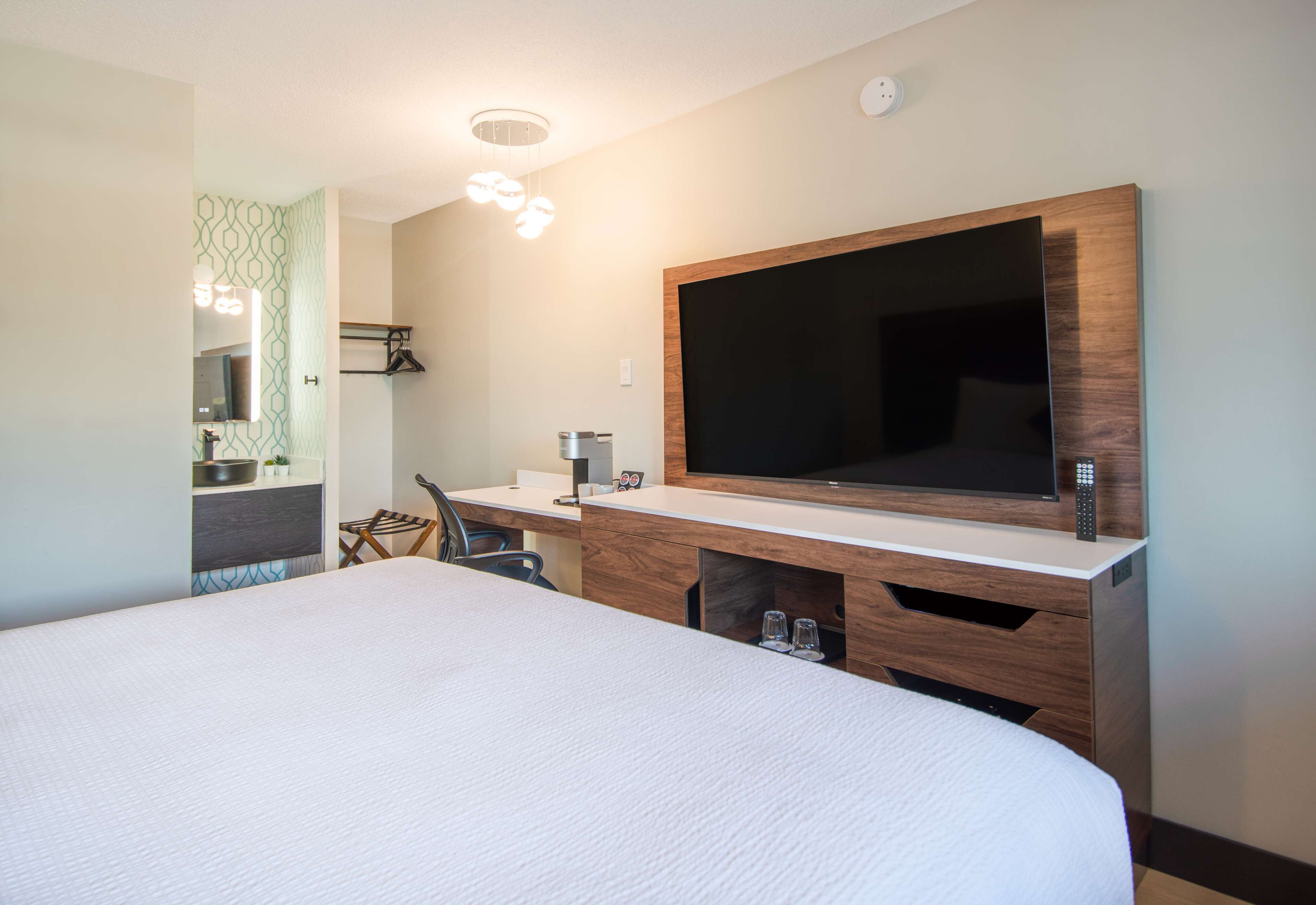 A hotel room with a large bed, a desk with a flat-screen TV, a mirror, and a small seating area.