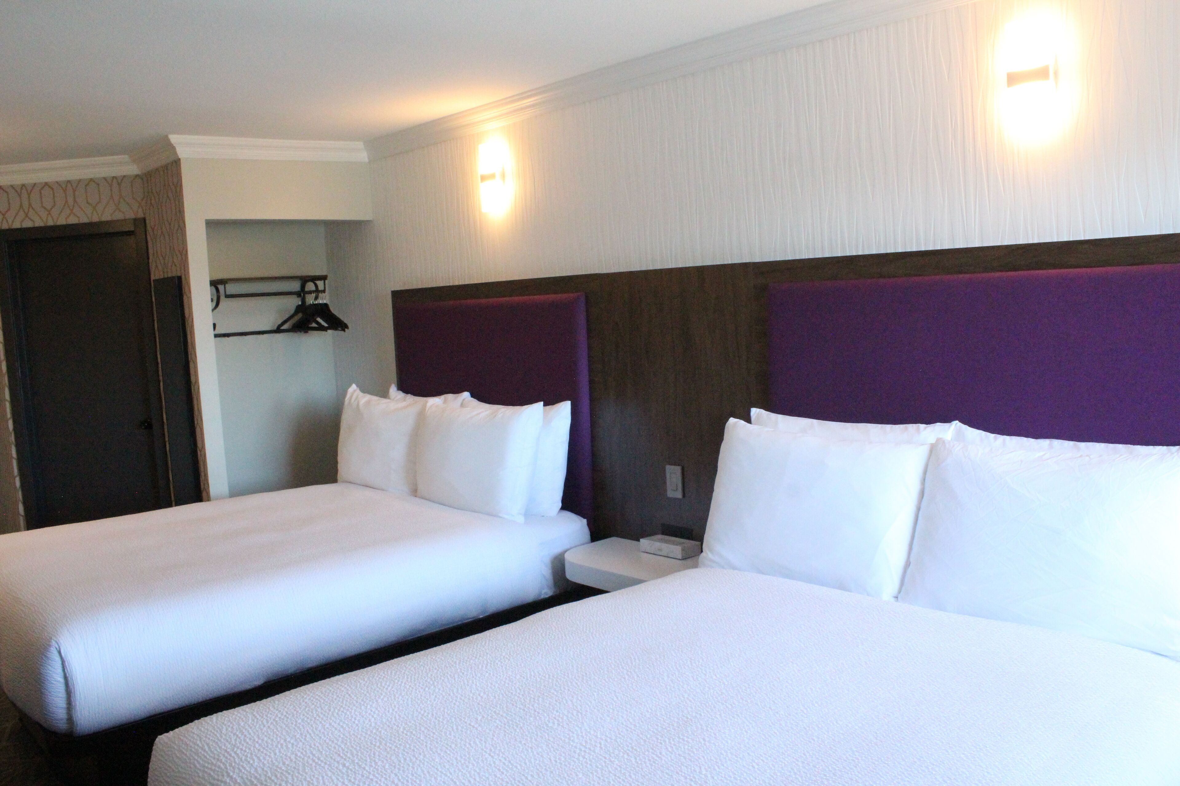 A hotel room with two single beds, a nightstand, a small shelf with a hanger, and a door.