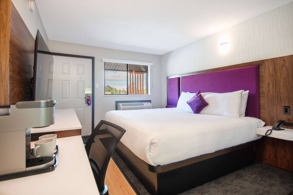 A hotel room with a bed, a desk with a coffee machine, and a window with blinds.