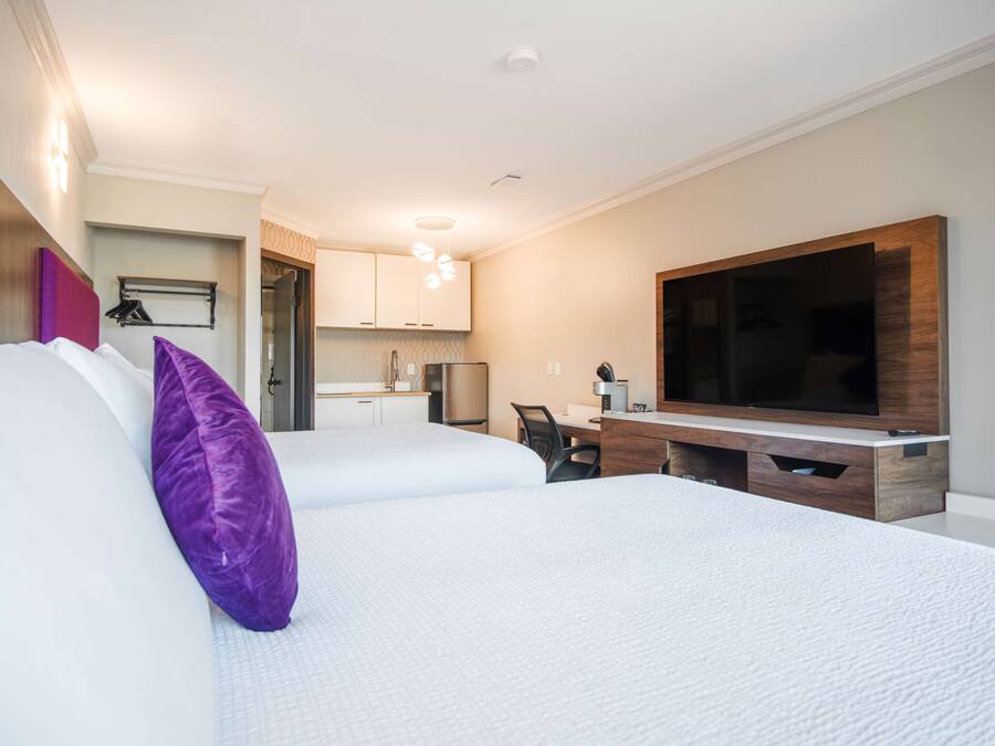 A hotel room with two beds, a TV, a desk, and a kitchenette.