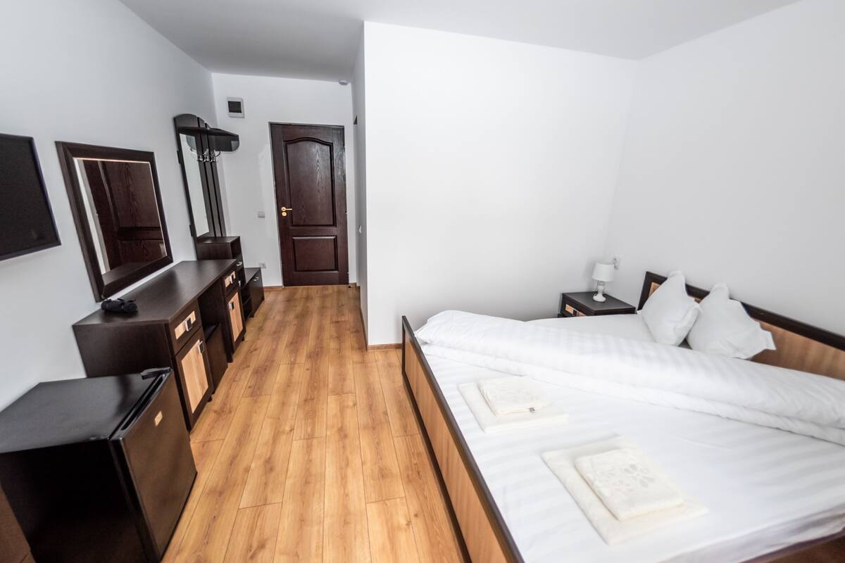 A hotel room with a wooden floor, a bed with white bedding, a small bedside table with a lamp, a wardrobe, and a mirror on the wall.