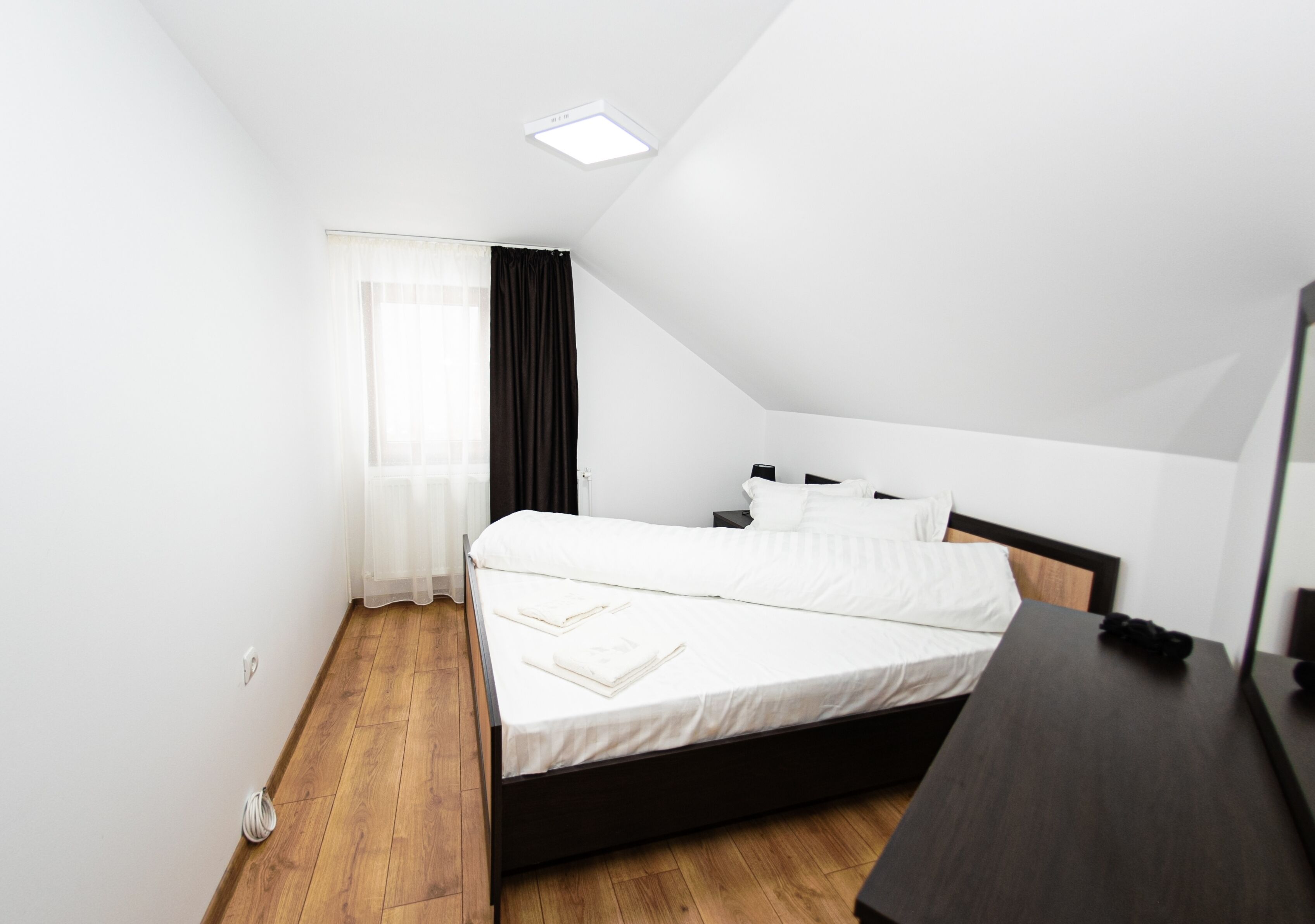 A modern bedroom with a wooden floor, a bed with white linens, a dark headboard, and a small bedside table.