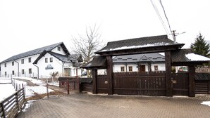 A traditional wooden gate with a snow-covered roof, a white building with a sign, and a paved pathway. - Casa Câmpulungeană (Campulung Moldovenesc)