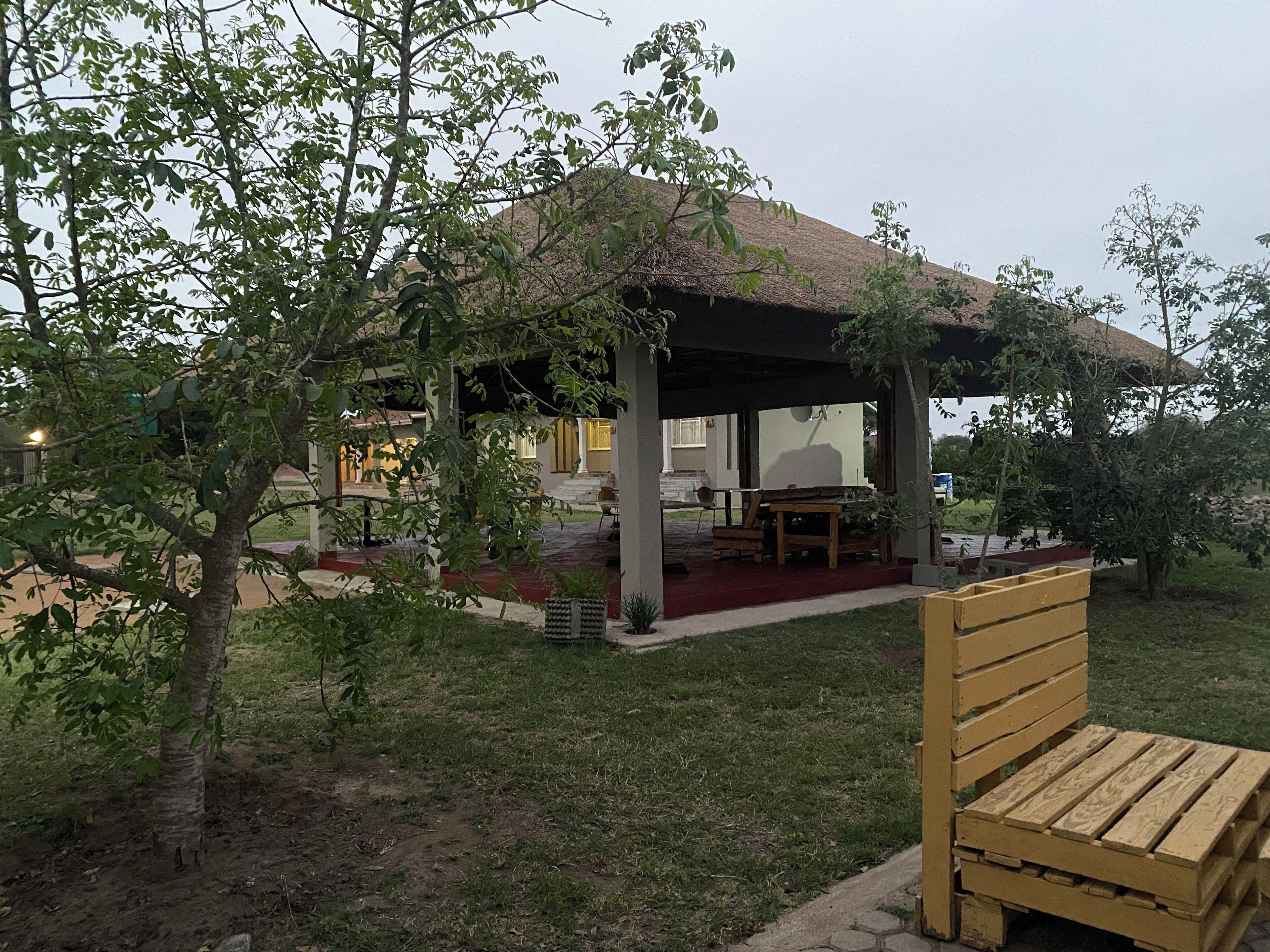 A covered outdoor seating area with a table and chairs, surrounded by greenery and a building in the background.