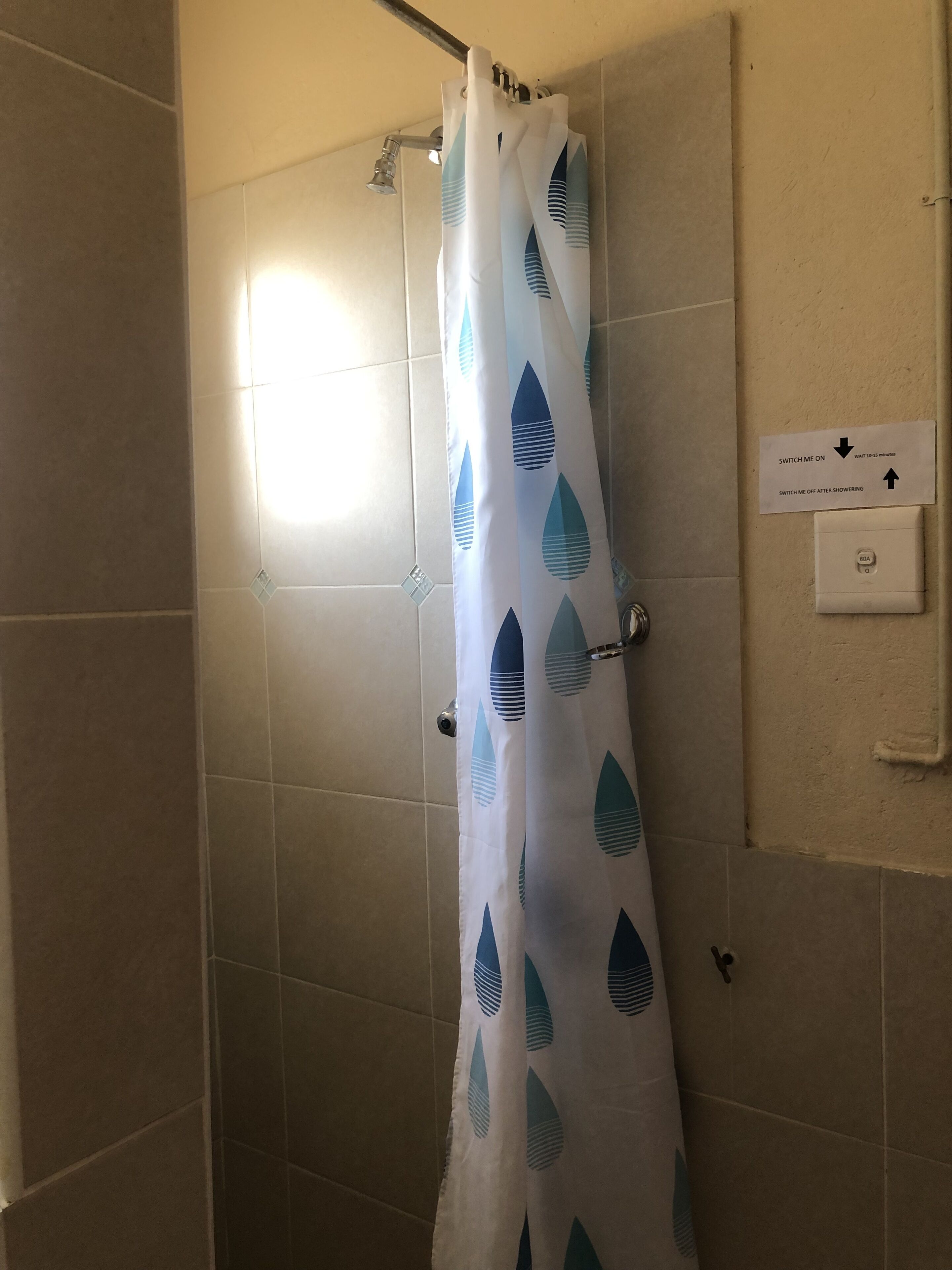 A shower area with a white curtain featuring blue leaf patterns, a showerhead, and a sign indicating a 10-15 minute wait time for the shower.