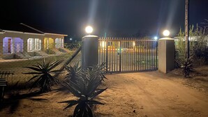 A gated entrance with a metal fence and two illuminated bollards, leading to a building with multiple lit windows. - Wanakah Estate Guesthouse (Mpaka)