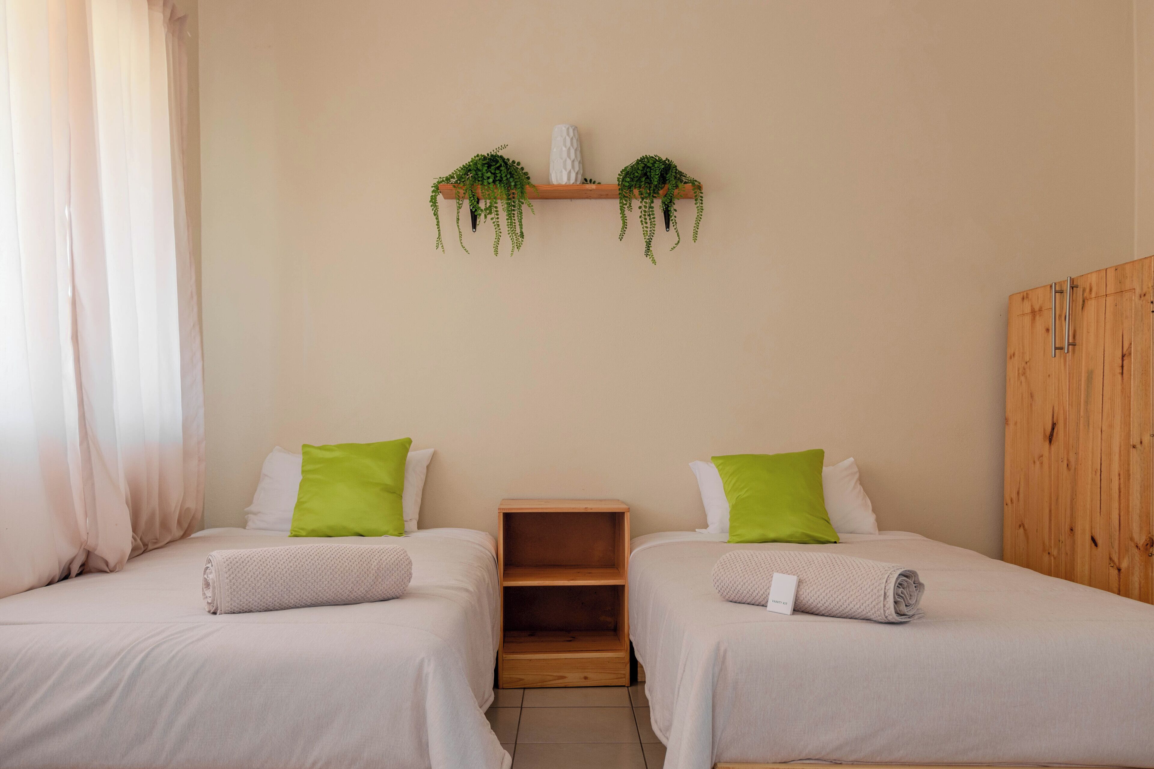 A room with two beds, each with a green pillow and a rolled towel. There is a wooden shelf with plants above the beds.