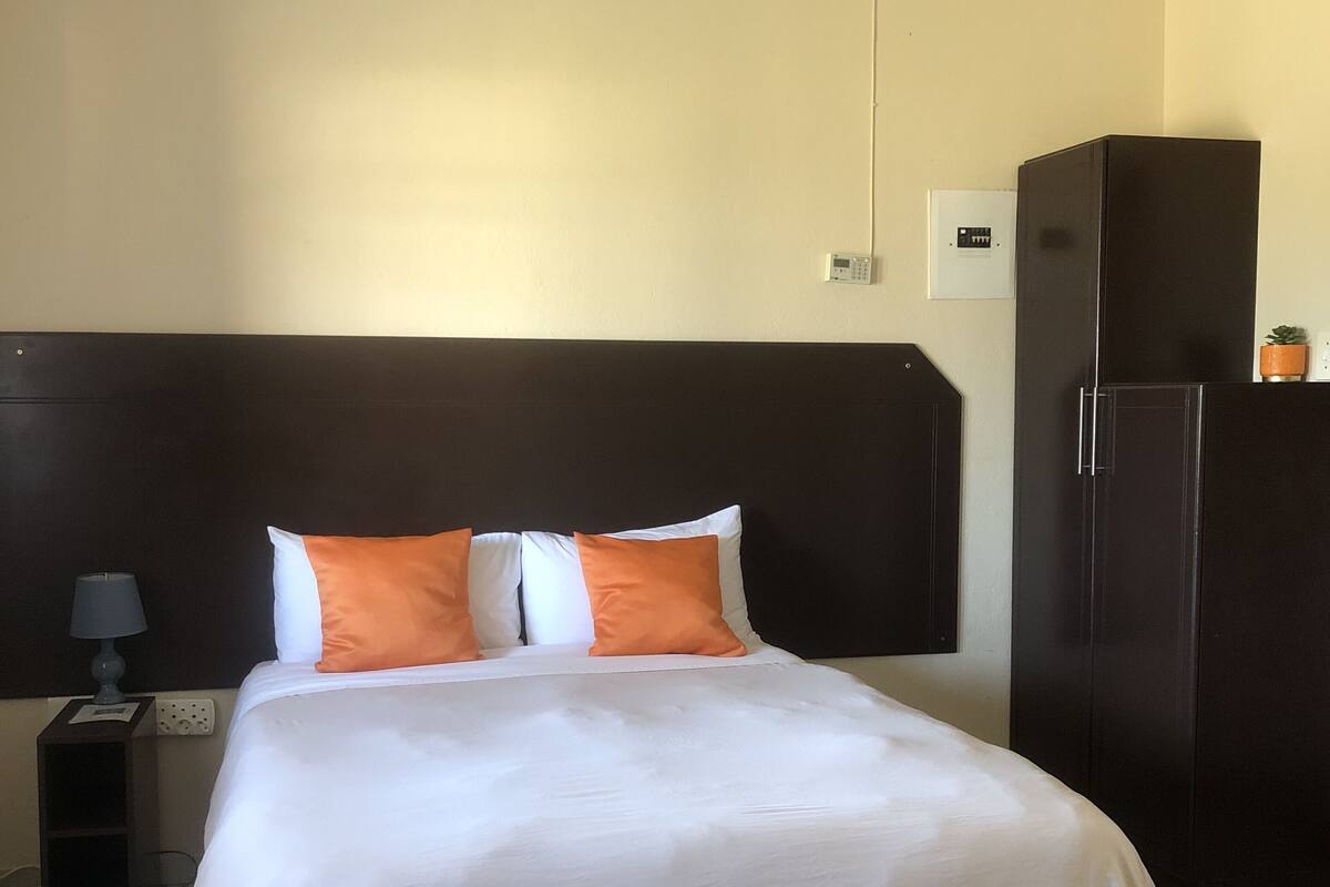 A hotel room with a bed, a nightstand, a lamp, a thermostat, and a refrigerator.