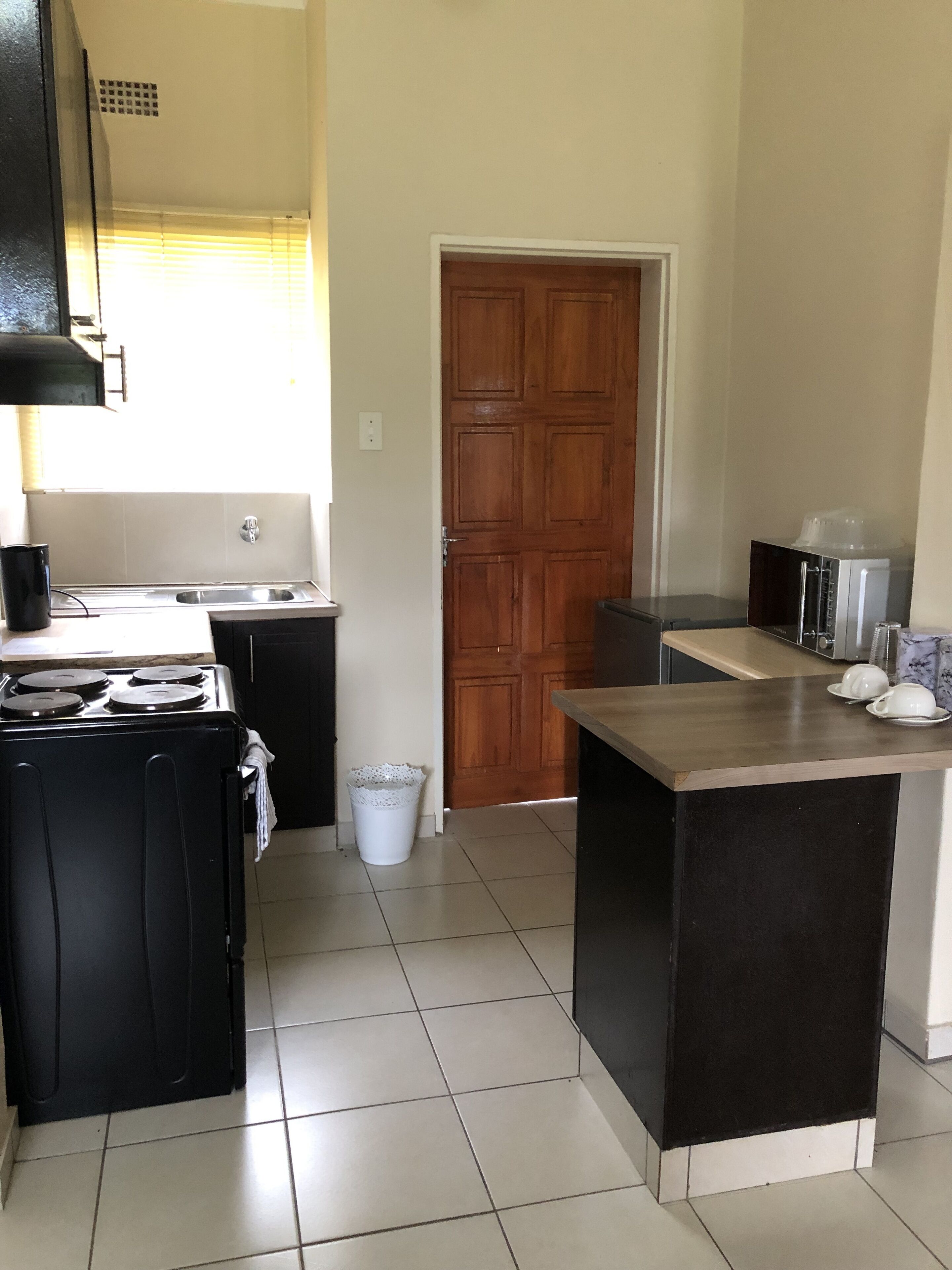 A compact kitchen with a stove, sink, and a microwave. There's a wooden door and a trash bin.