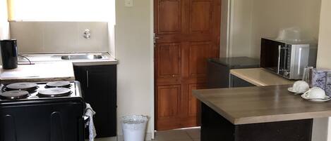 A compact kitchen with a stove, sink, and a microwave. There's a wooden door and a trash bin.