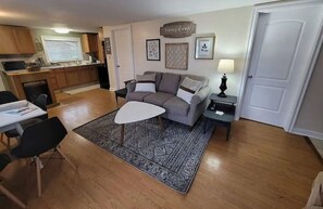 Sala de estar com sofá, mesa de centro e tapete. Há uma área de jantar com cadeiras e mesa. O cômodo possui piso de madeira e uma cozinha ao fundo.