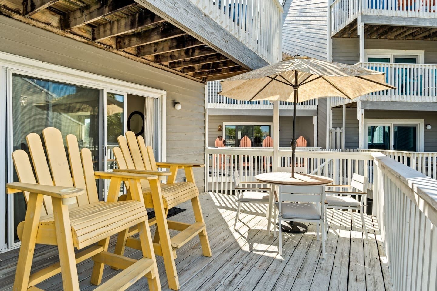 A wooden deck with Adirondack chairs and a table, a white umbrella, and a balcony with railings.