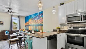 Una cucina moderna con mobili bianchi, lavastoviglie integrata, doppio forno e microonde. C'è una zona pranzo con tavolo e sedie, e un grande quadro che raffigura una veduta cittadina sulla parete.