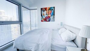 Een moderne hotelkamer met een groot raam, een bed met witte lakens, een nachtkastje met een lamp en een kleurrijk schilderij aan de muur.