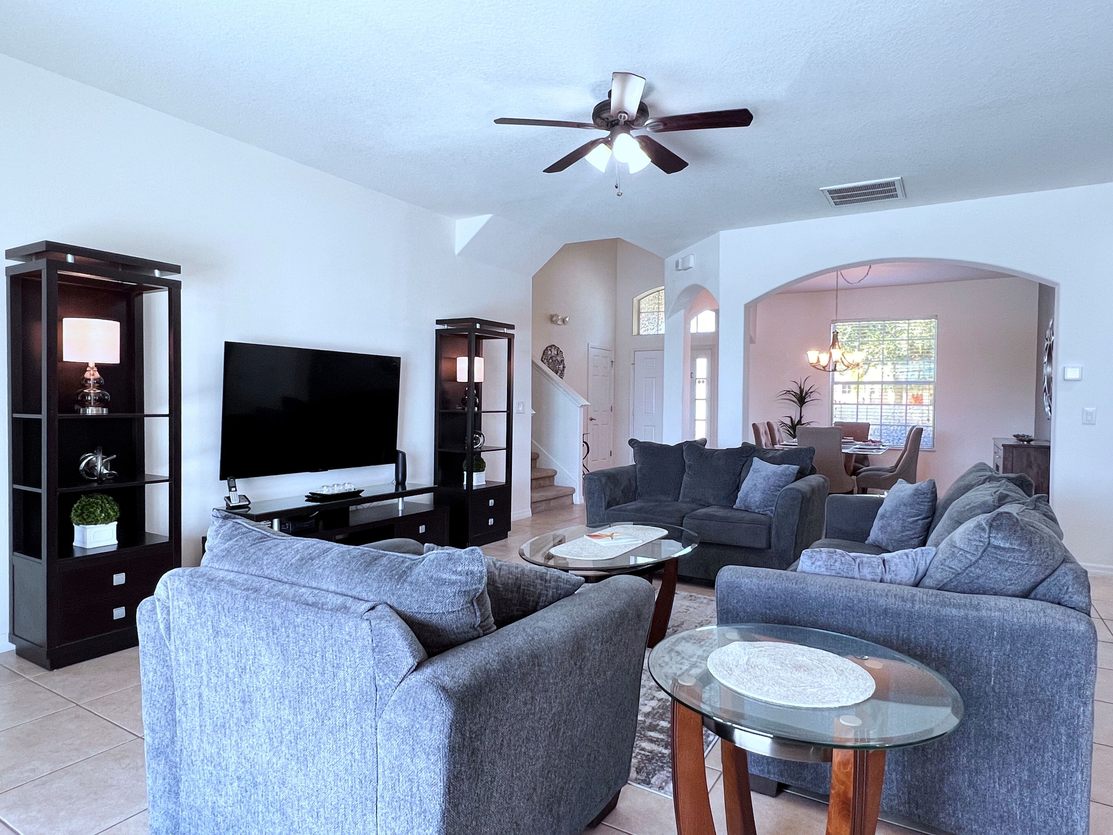 A spacious living room with a flat-screen TV, grey sofas, a glass coffee table, and a ceiling fan.