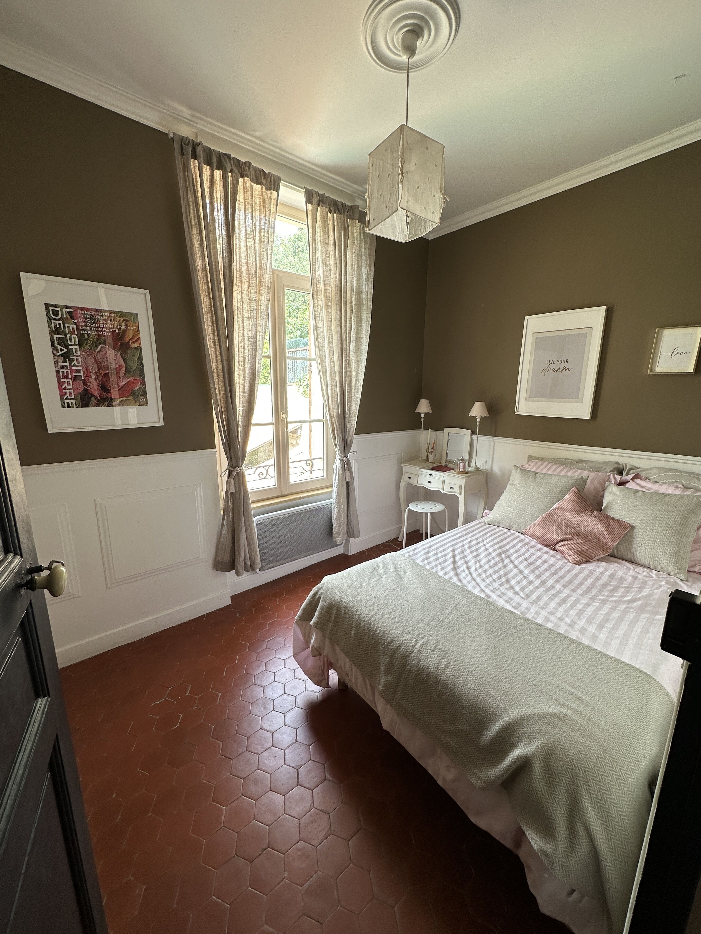 Une chambre à coucher avec un lit, une fenêtre avec des rideaux, une table de chevet et un luminaire fixé au mur.