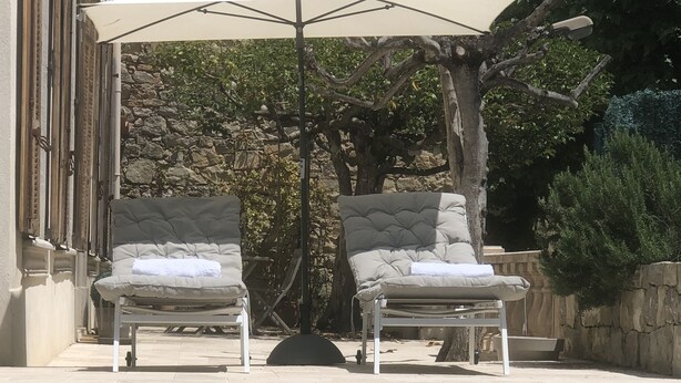 Zona de piscina con dos sillones de descanso bajo una pérgola, un muro de piedra y un árbol.