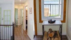 A hallway with a wooden floor, a staircase with a black railing, and a window overlooking a body of water.