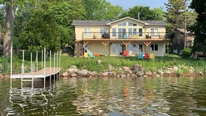 A two-story house with a dock extending into a lake, surrounded by greenery.