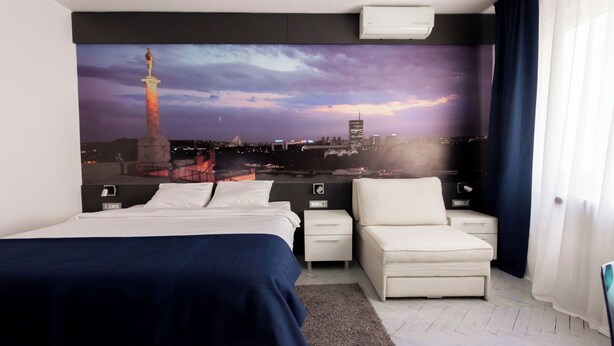 Habitación de hotel con cama, una silla, mesita de noche, aire acondicionado instalado en la pared y un gran mural con vista a la ciudad.
