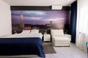 A hotel room with a bed, a chair, a nightstand, a wall-mounted air conditioner, and a large cityscape mural.