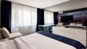 Habitación de hotel con cama, escritorio, silla, televisor y un amplio ventanal con cortinas.