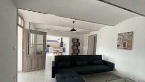 A modern living room with a dark sofa, a glass coffee table, and a kitchen in the background. - Gîte « Les Oliviers » Dans Mas Typiquement Ardéchois - 6 Personnes (Saint-Sauveur-de-Cruzières)